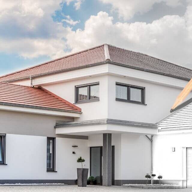 Maison moderne à toit en pente, avec façade blanche et fenêtres grises sous un ciel nuageux.